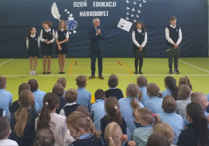 Dyrektor na tle uczniów występujących i dekoracji składa życzenia z okazji Dnia Edukacji Narodowej